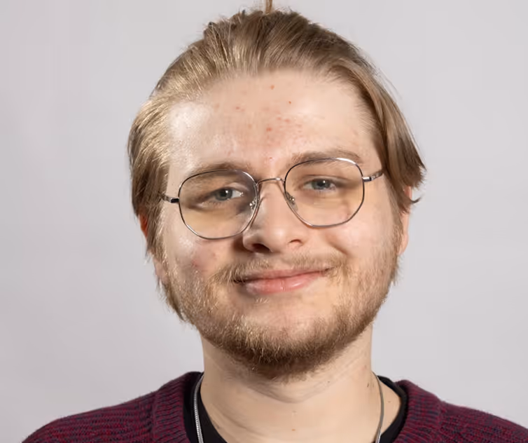 Smiling young man with glasses, light brown hair pulled back, and a short beard wearing a maroon sweater.