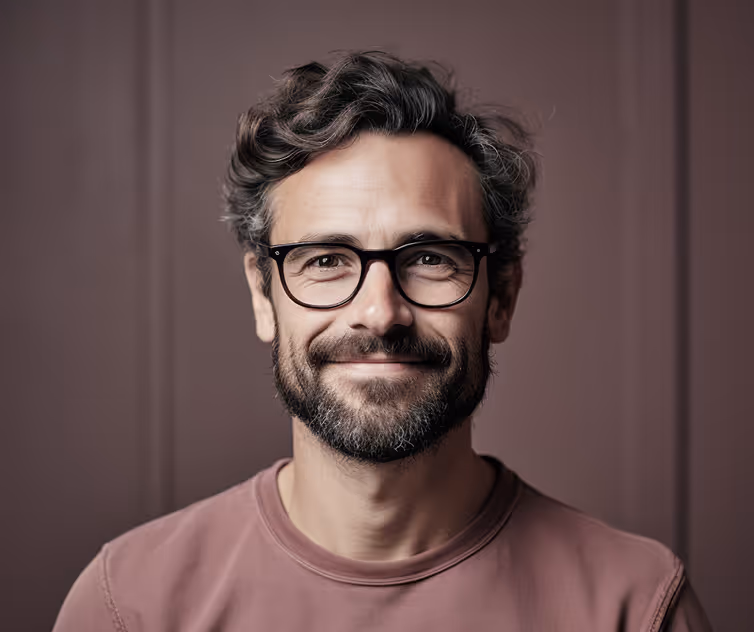 Smiling man with curly hair, beard, and glasses wearing a mauve sweatshirt against a mauve background.