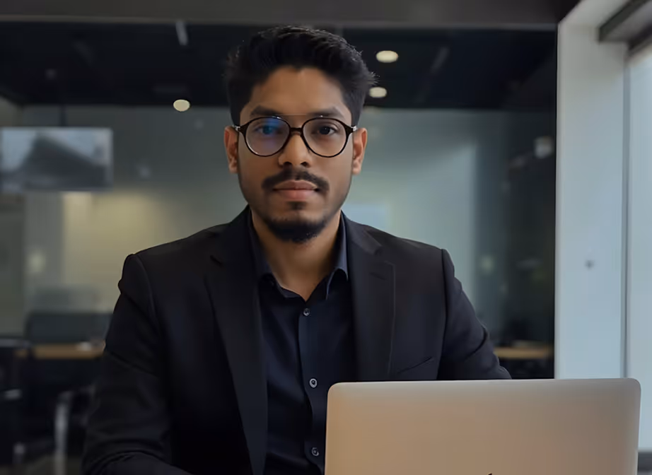 Man wearing glasses and a black suit sitting behind a laptop in an office setting.