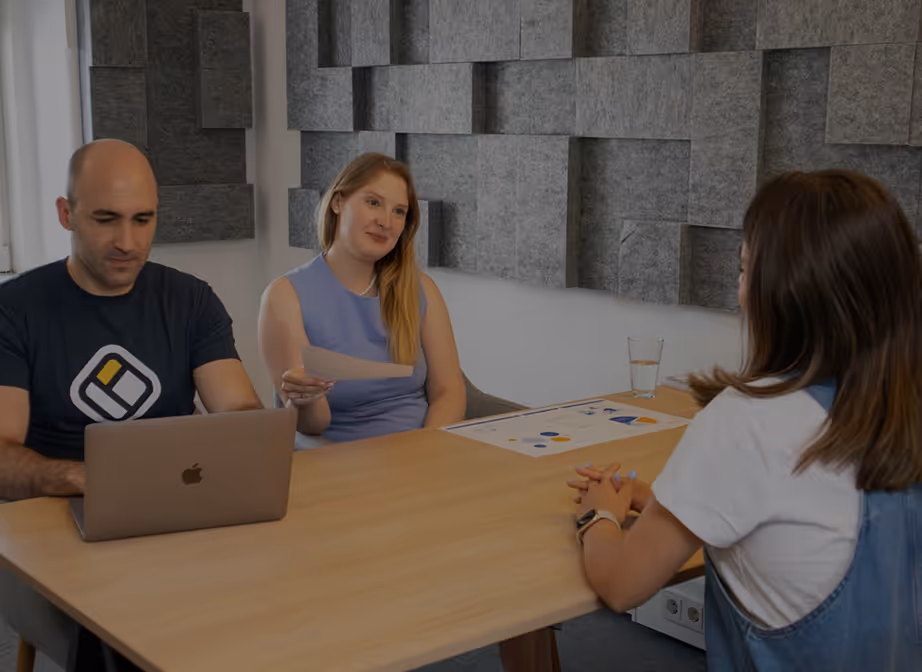 A man and a woman sitting at a table interviewing another woman across from them, with charts and a glass of water on the table.