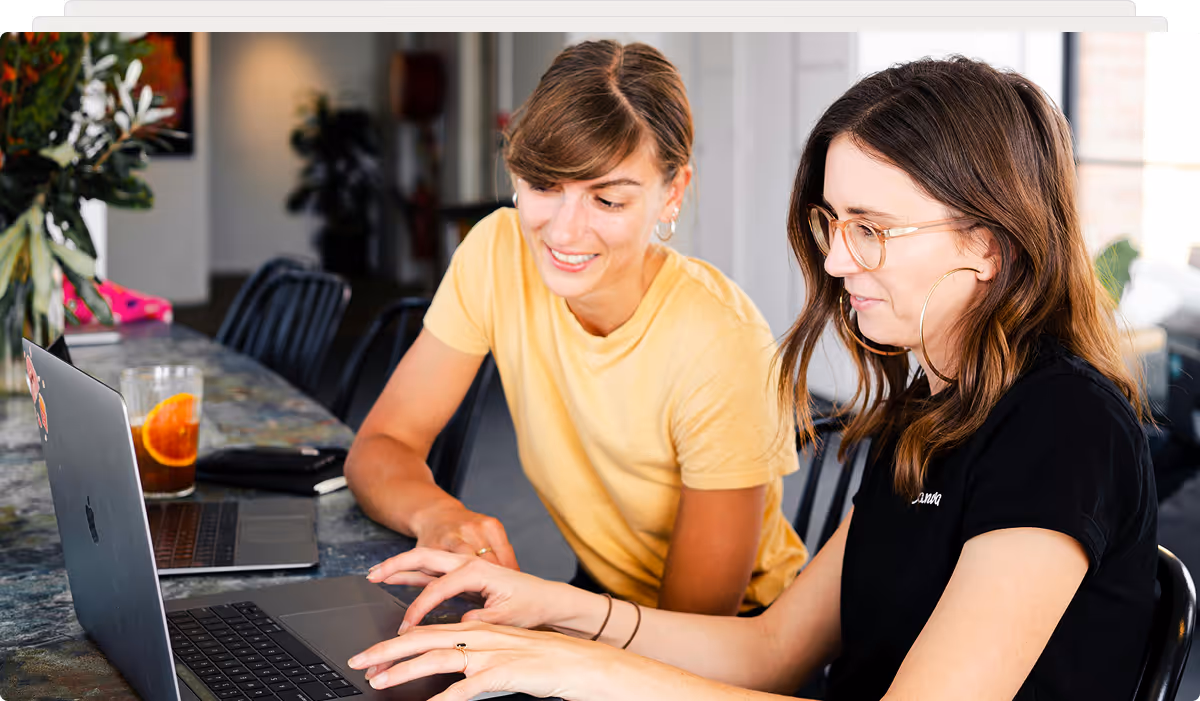 Two women collaborating and working together on a laptop at a table with a drink and a second laptop nearby.