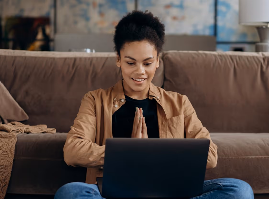 Smiling woman sitting cross-legged on the floor in front of a couch, with hands pressed together, looking at a laptop.