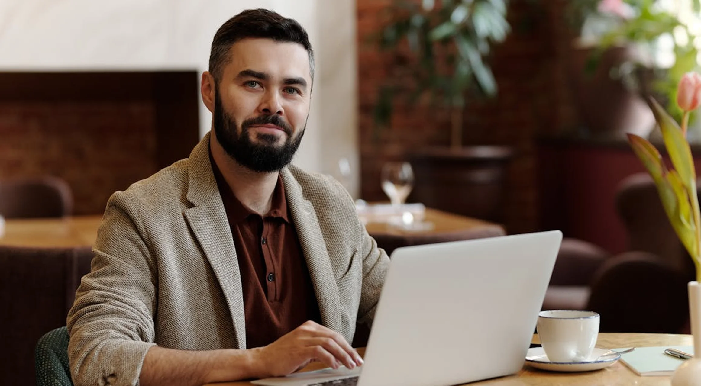 Smiling man with beard working on a laptop in a cozy cafe setting with a cup and flowers on the table.